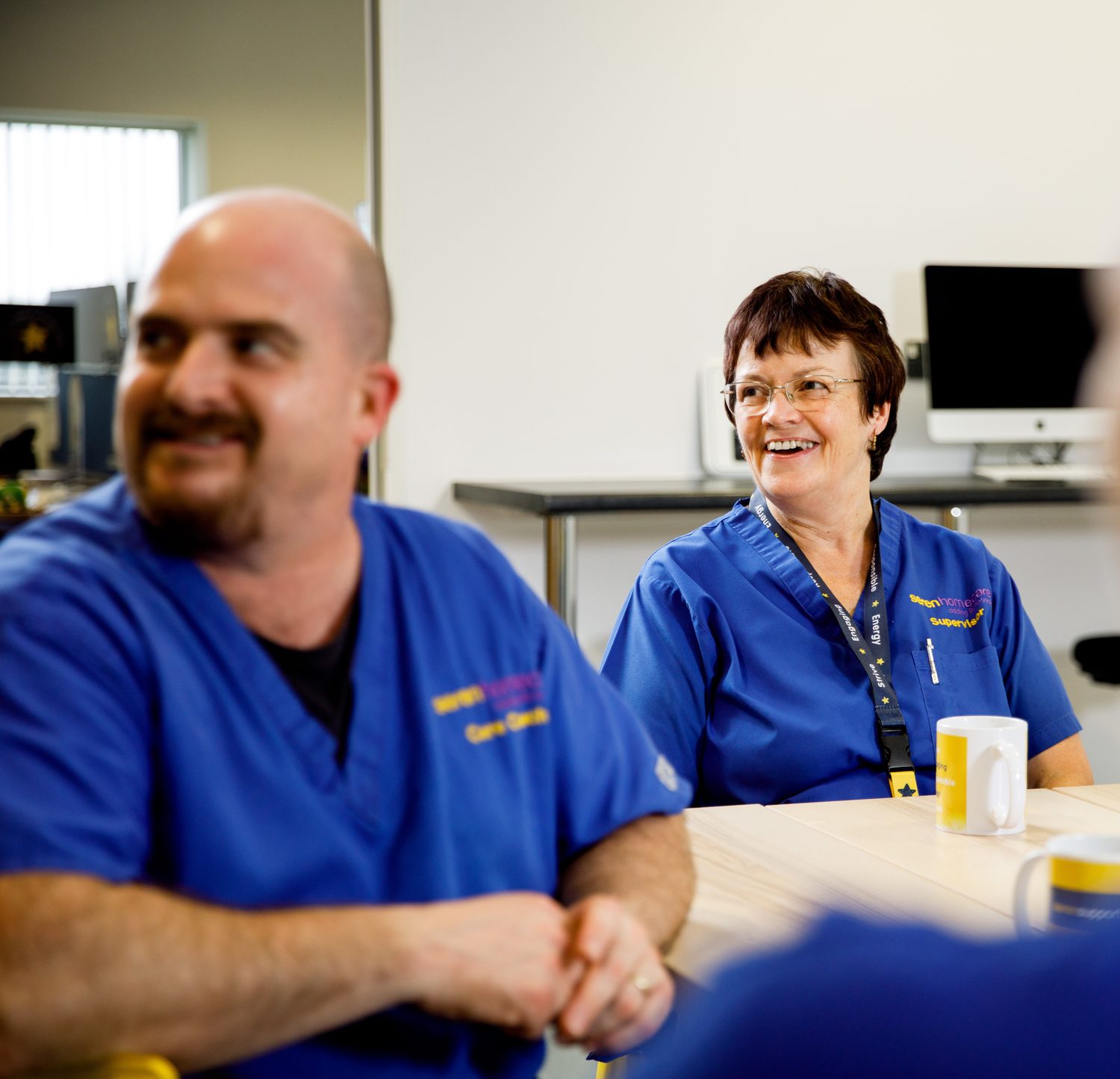 Two Seren Support Services care staff in uniform sitting round a table, talking and laughing during a team catch-up.